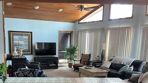Un salón espacioso con ventilador de techo, ventanas grandes con persianas, un televisor de pantalla plana y una zona de estar cómoda con sofá y sillones.