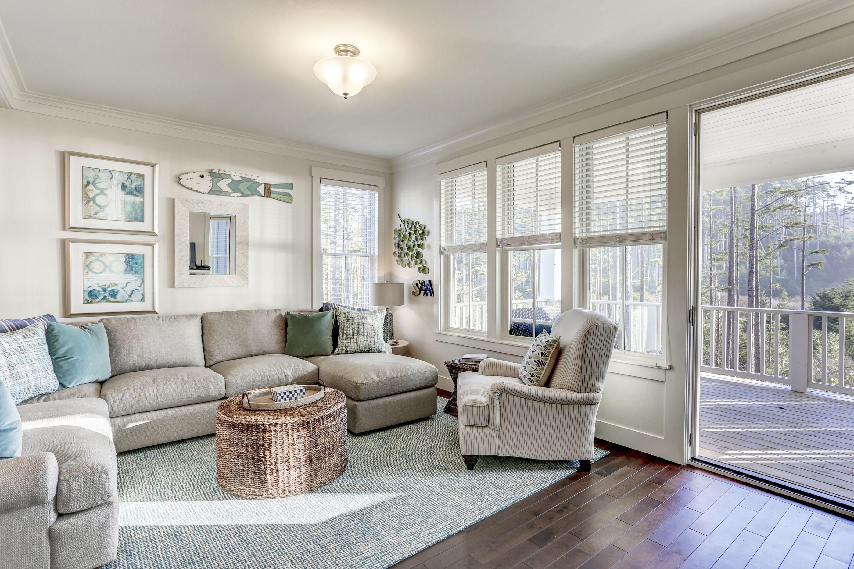 A spacious living room with a large gray sectional sofa, a striped armchair, and a wicker ottoman. There are multiple windows with blinds, a ceiling light, and framed artwork on the walls.