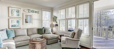 A spacious living room with a large gray sectional sofa, a striped armchair, and a wicker ottoman. There are multiple windows with blinds, a ceiling light, and framed artwork on the walls.