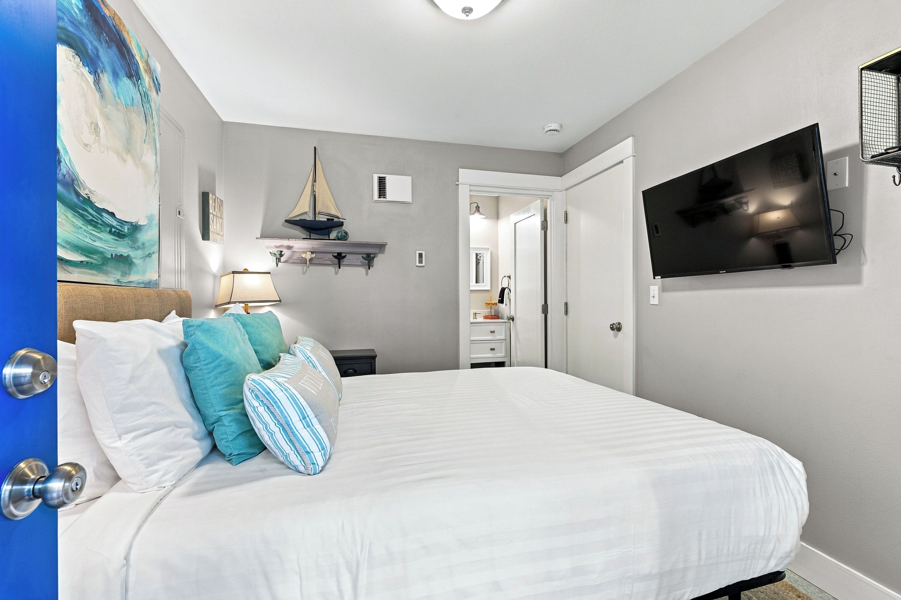 Un dormitorio con cama, televisor montado en la pared, una pequeña estantería con objetos decorativos y una puerta que da a otra habitación.
