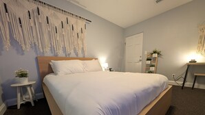 Een net opgemaakt bed in een kamer met een macramé wanddecoratie, een klein rond tafeltje met een plant en een houten nachtkastje met een lamp.