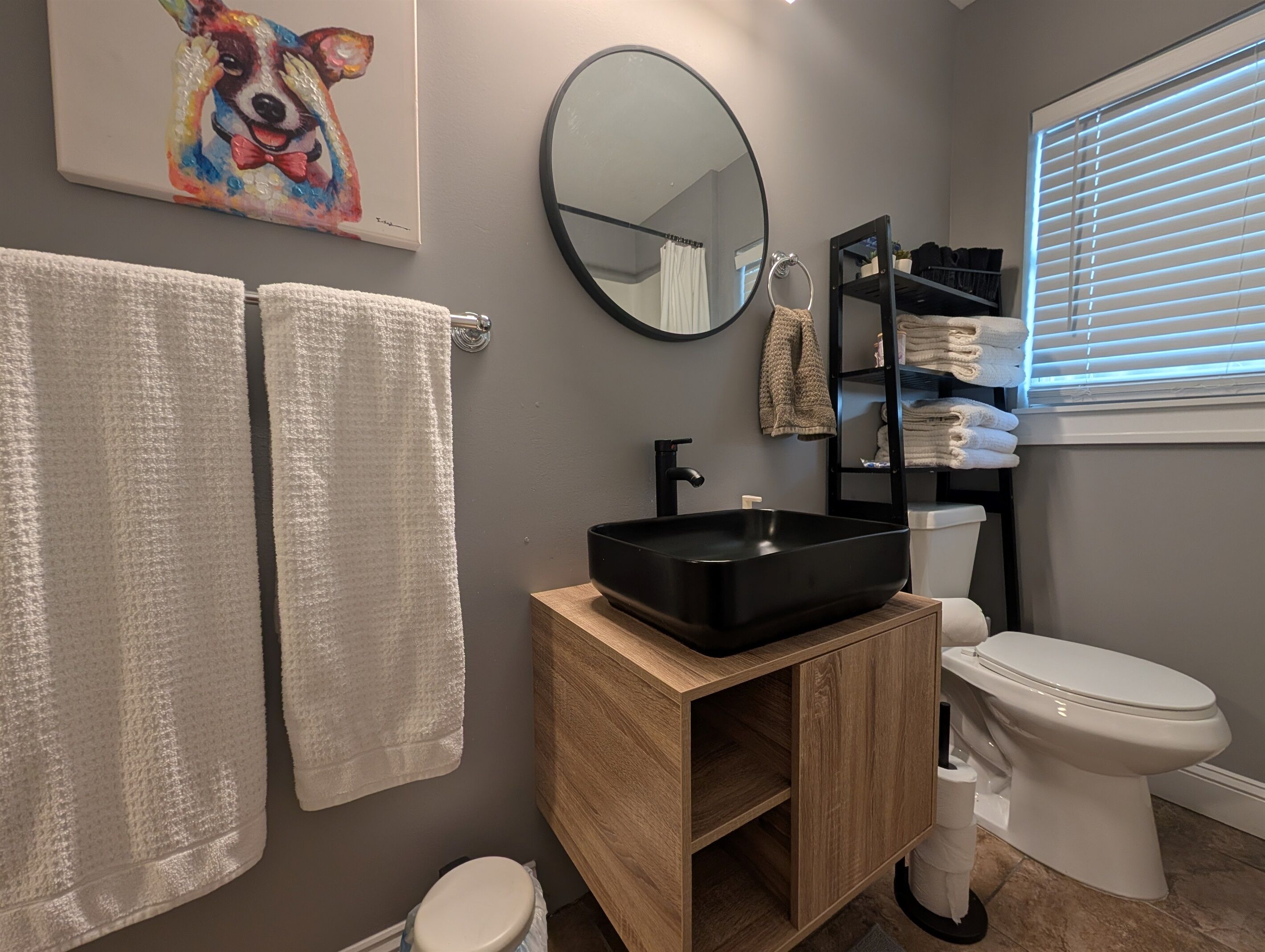 A modern bathroom with a wooden vanity, black sink, and white toilet. There are two white towels hanging on the wall, a mirror, and a shelf with folded towels.