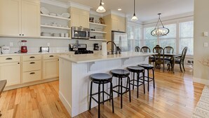 Une cuisine moderne avec des meubles blancs, un îlot central doté de tabourets de bar, des appareils en acier inoxydable et un coin repas avec des chaises en bois et un lustre.