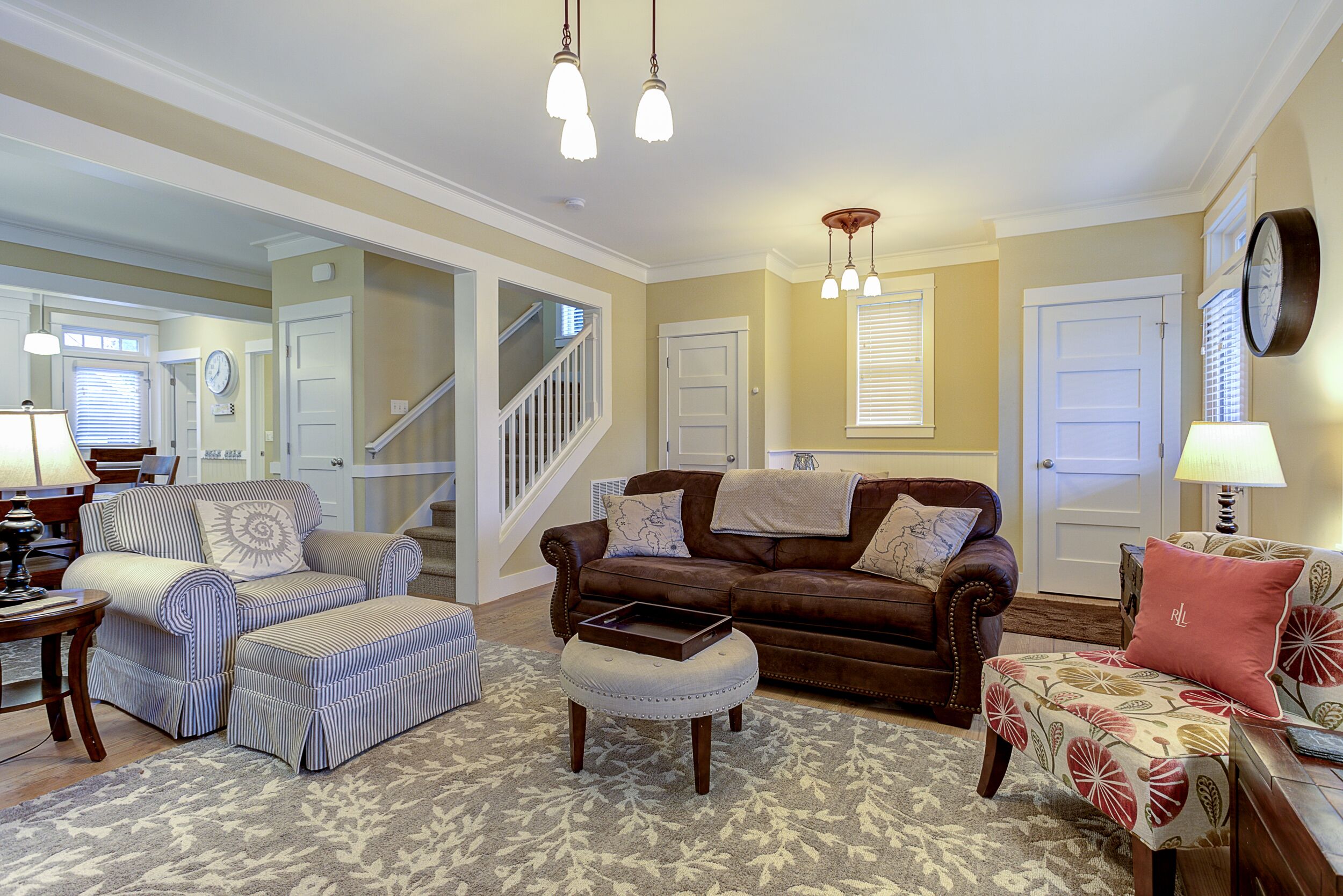 Ein Wohnzimmer mit braunem Ledersofa, gestreiften Sessel und Poufs, einem gemusterten Teppich und einer Treppe, die zu einer anderen Ebene führt.