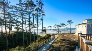 A coastal path leading to a beach with a wooden fence, surrounded by tall pine trees and a building with a balcony overlooking the ocean.