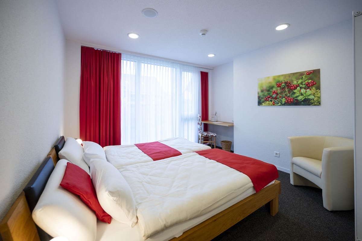 A neatly made bed with red and white bedding, a chair, and a painting on the wall.