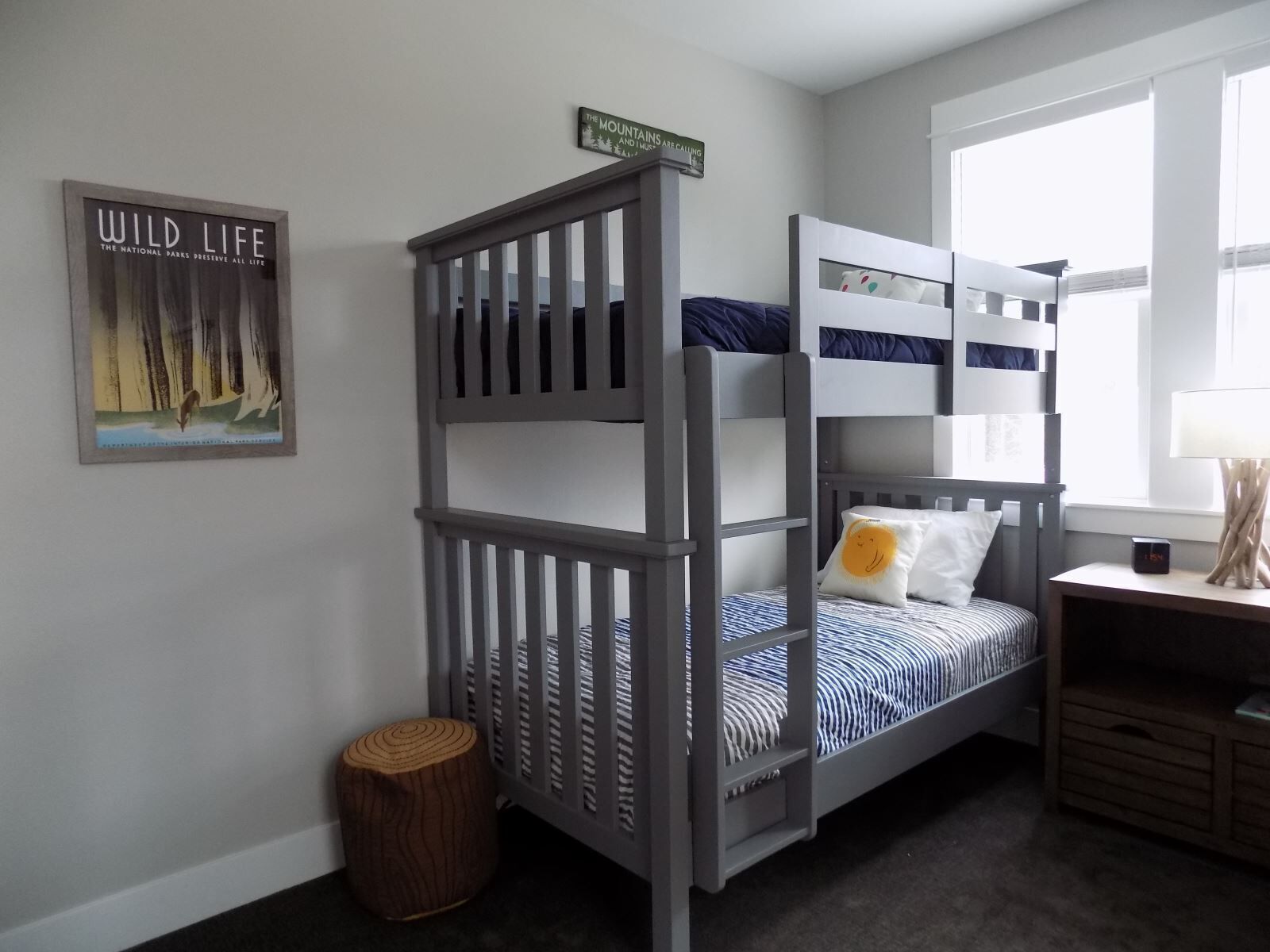 A child's bunk bed with a grey frame, a small round stool, a framed poster, and a window with natural light.