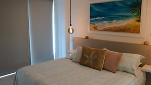 Et soveværelse med en seng, puder, et billede af en strand og en hængende lampe.