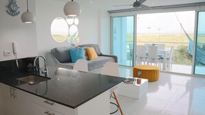 Et moderne køkken med en central ø, en grå sofa med farverige puder og en stor skydedør, der fører ud til et udeområde med havudsigt.