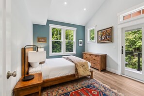 A bedroom with a bed, bedside table, lamp, dresser, and large windows.
