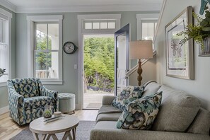 A living room with a sofa, armchair, and a lamp. There is a door leading to a balcony with a view of trees. - Seabrook Creekside Cottage | Hot Tub, Location 3BR | Sleeps 6, South Farm (Pacific Beach)