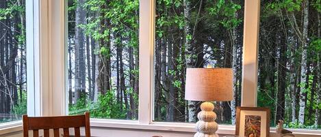 A room with a large window offering a view of a forest. There is a wooden chair with a cushion, a small wooden table, and a wooden dresser with drawers. A lamp with a white shade is on the dresser.