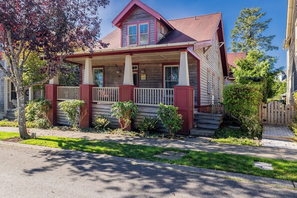 A two-story house with a porch, a red roof, and a small front yard with a tree and shrubs.