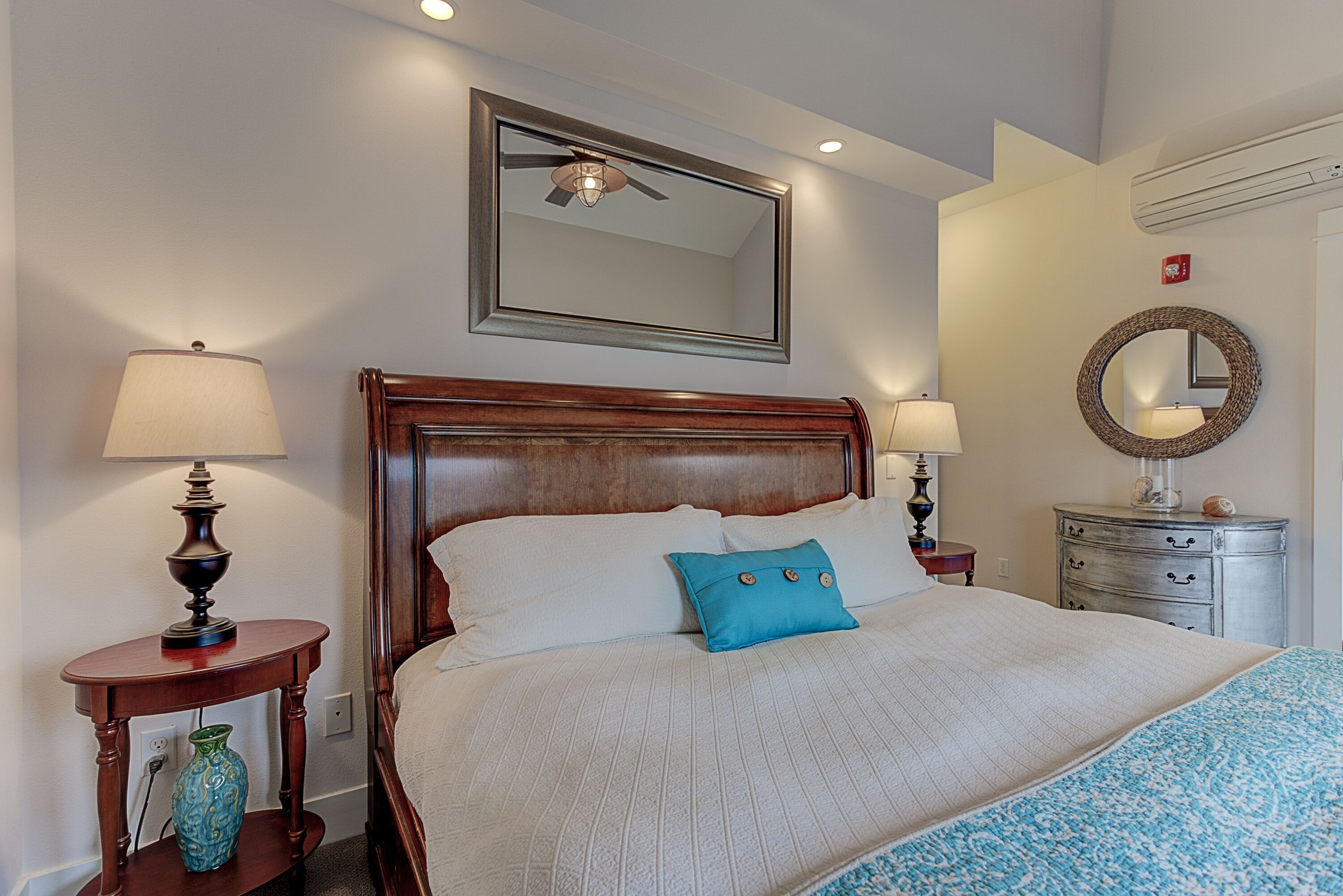 A neatly made bed with a wooden headboard, two bedside tables with lamps, a round mirror, and a small dresser with drawers.