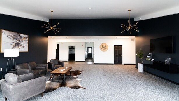 A modern living room with a grey sofa, a coffee table, and a rug. There is a fireplace, a TV mounted on the wall, and a chandelier. - Conestoga Quarters (Jefferson City)