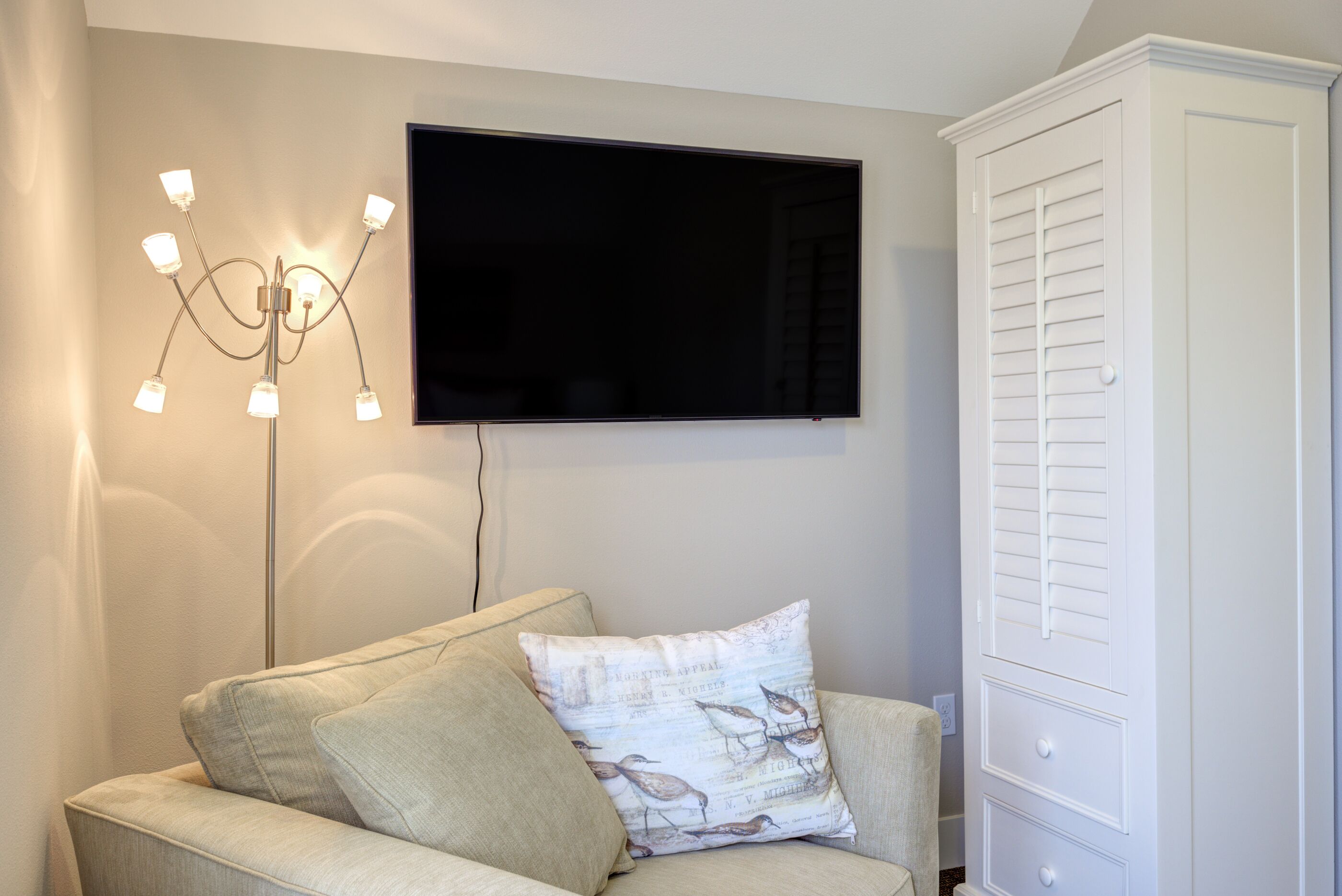 Ein Zimmer mit einem an der Wand befestigten Flachbildfernseher, einer Leuchte mit mehreren Lampen, einem Sofa mit einem gemusterten Kissen und einem weißen Kleiderschrank.