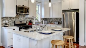 Une cuisine moderne avec des meubles blancs, des appareils en acier inoxydable, un grand îlot central doté de tabourets et un éclairage suspendu.