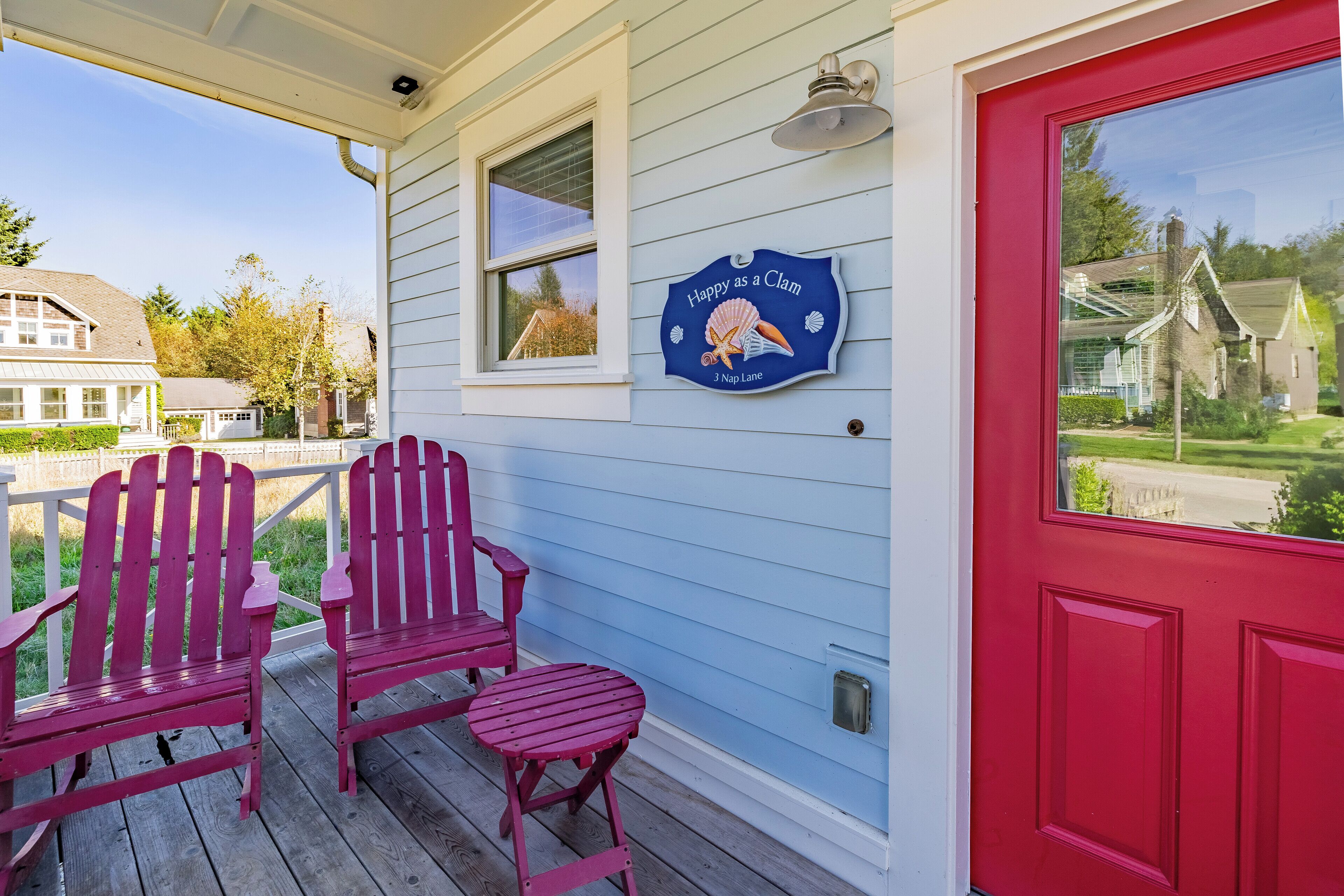 A porch with two Adirondack chairs and a small table, a red door, and a blue sign with a clamshell design.