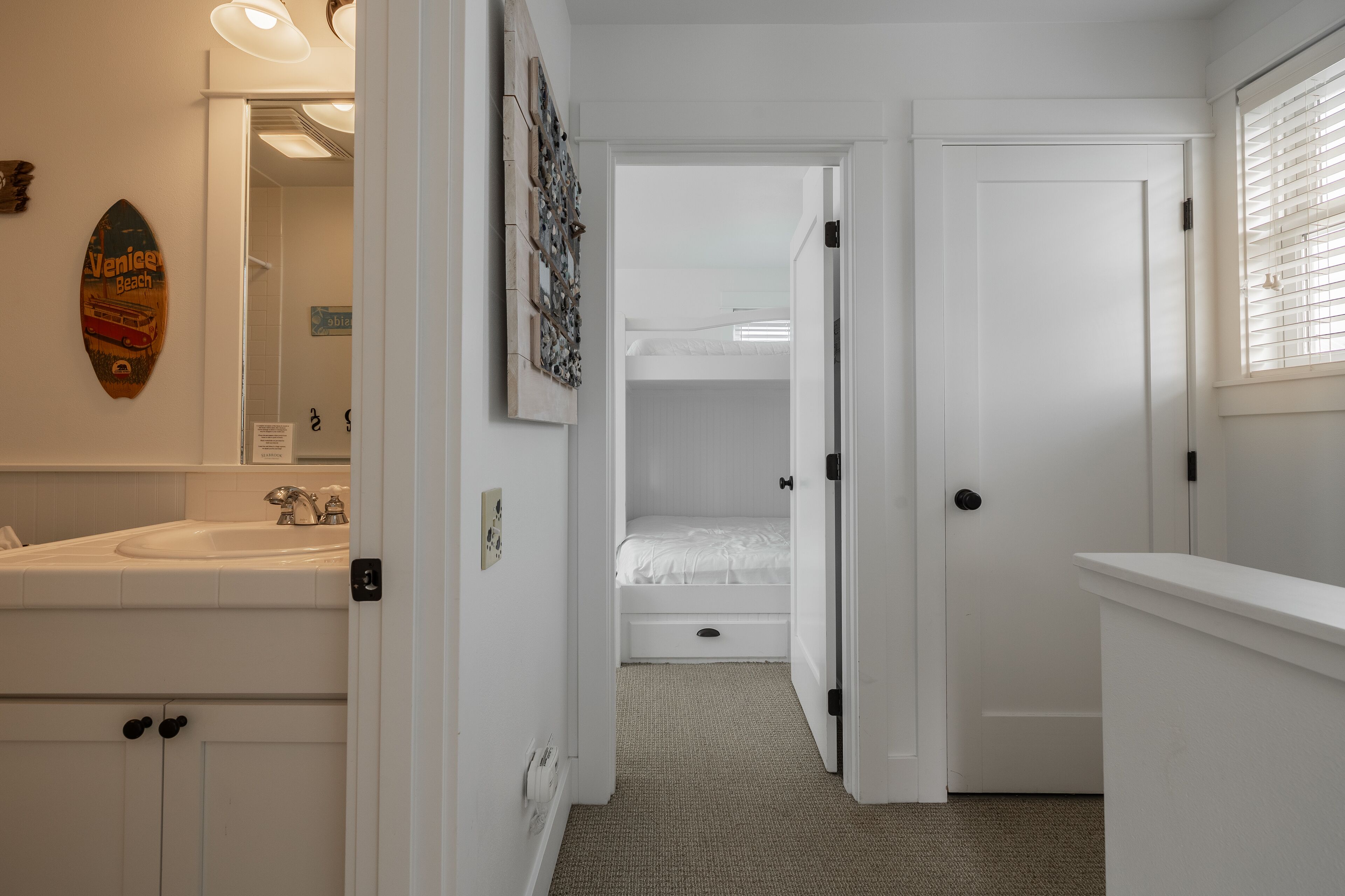A narrow hallway leading to a bedroom with a bunk bed, a bathroom with a sink and mirror, and a door with a window.