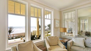 Ein Wohnzimmer mit großem Fenster und Blick auf einen Strand, zwei Sessel, ein Sofa und eine Lampe.