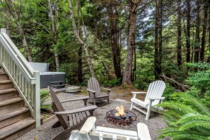 A forested area with a fire pit, Adirondack chairs, a wooden deck, and a hot tub.