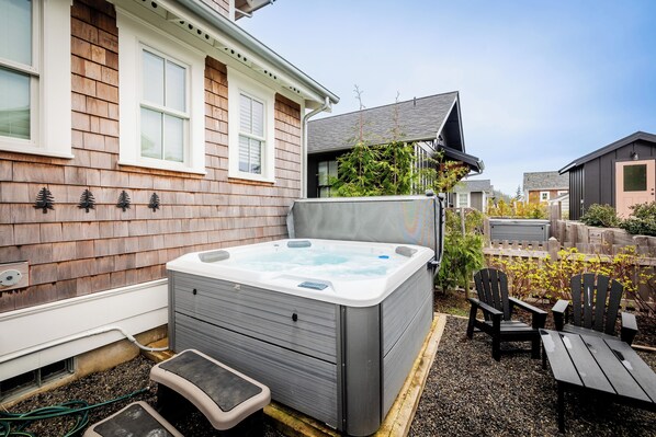 A hot tub in a backyard with a wooden house, a small deck, and two Adirondack chairs.