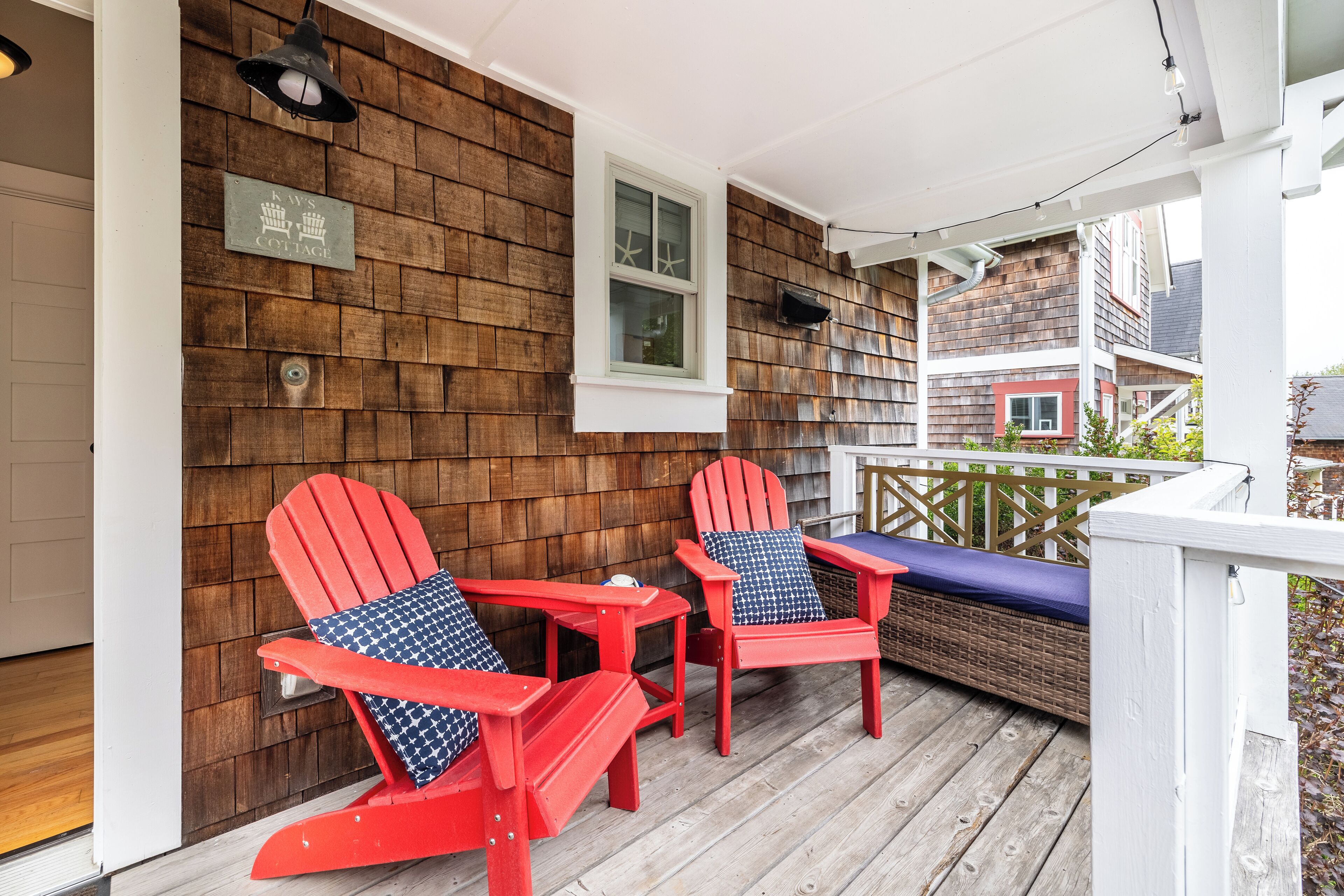 Ett trädäck med två röda Adirondack-stolar och ett litet bord, en trävägg med ett fönster och en skylt med texten "Kays Cottage".