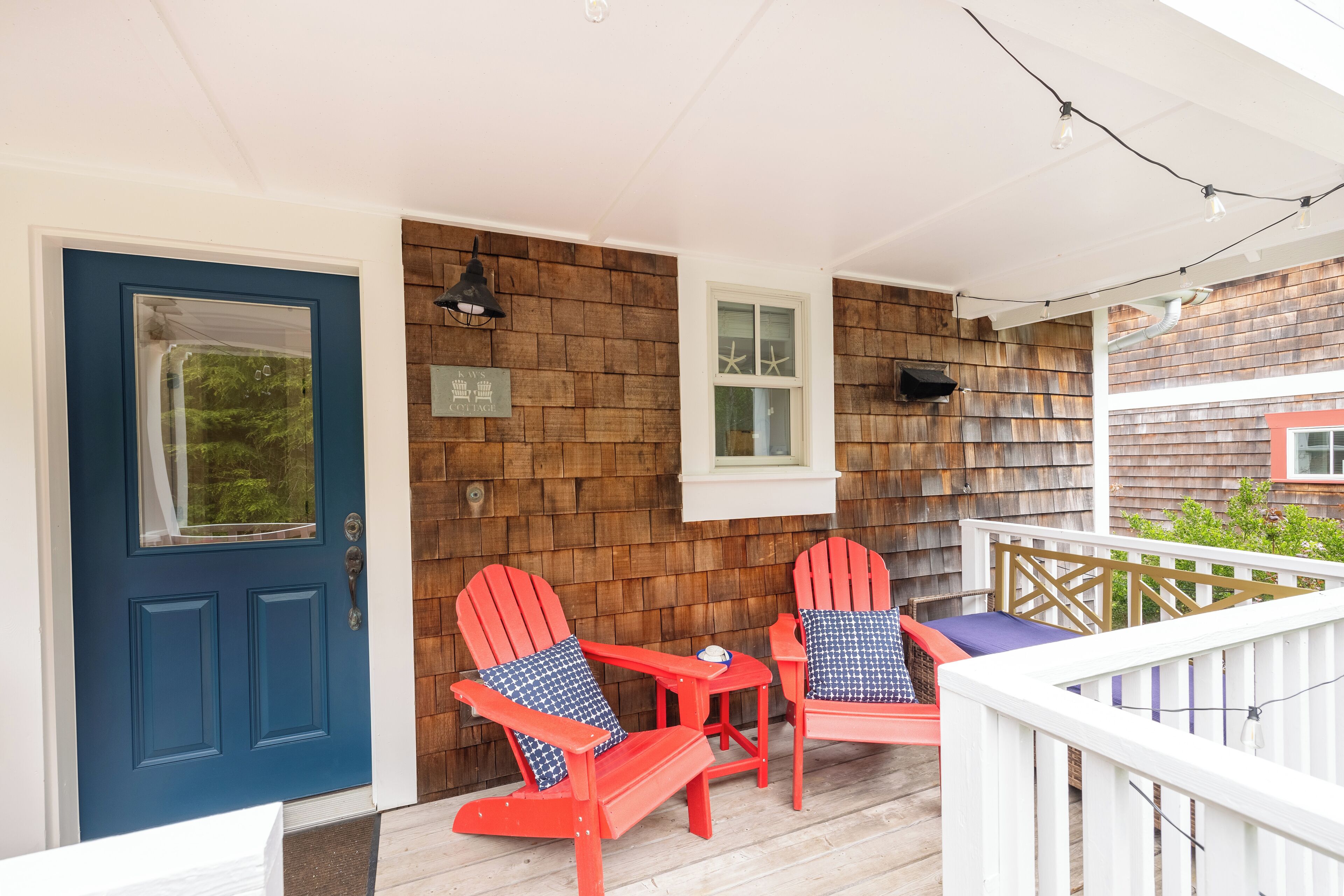 A wooden deck with two red Adirondack chairs and a small table, a blue door, and a white railing.