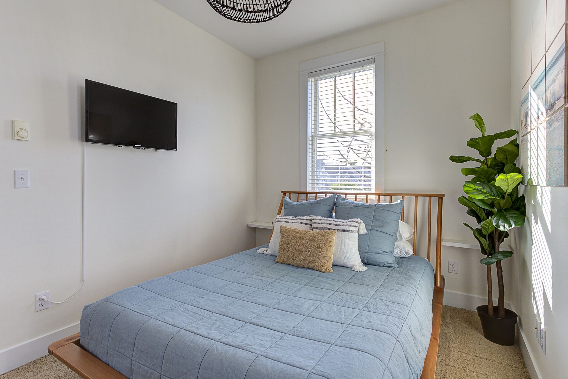 Een slaapkamer met een bed, een televisie aan de muur, een raam met zonwering en een kamerplant.
