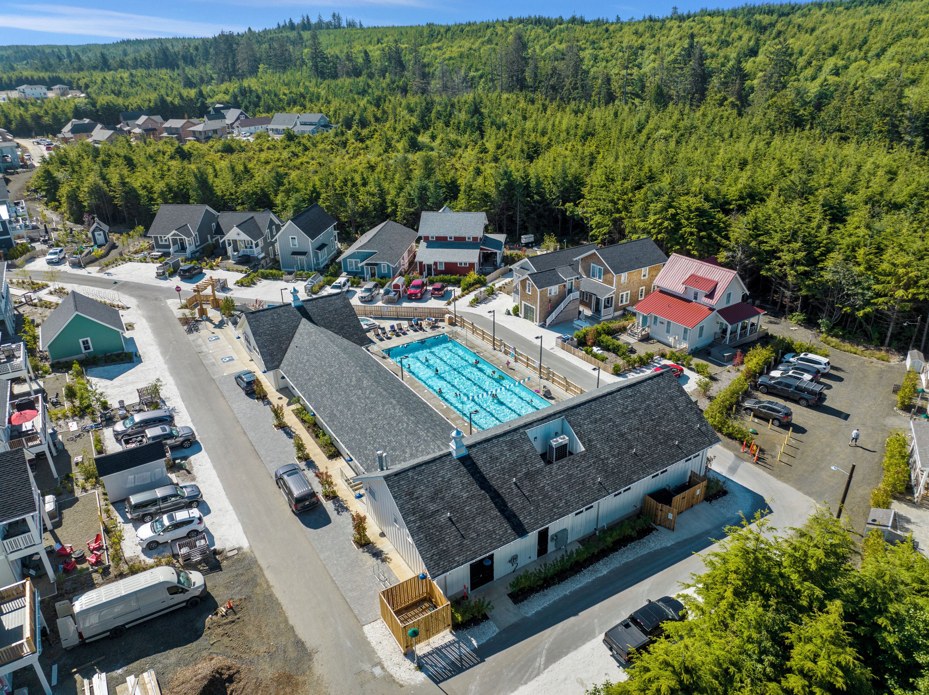 An aerial view of a residential area with a large swimming pool surrounded by houses, a road, and parked cars.