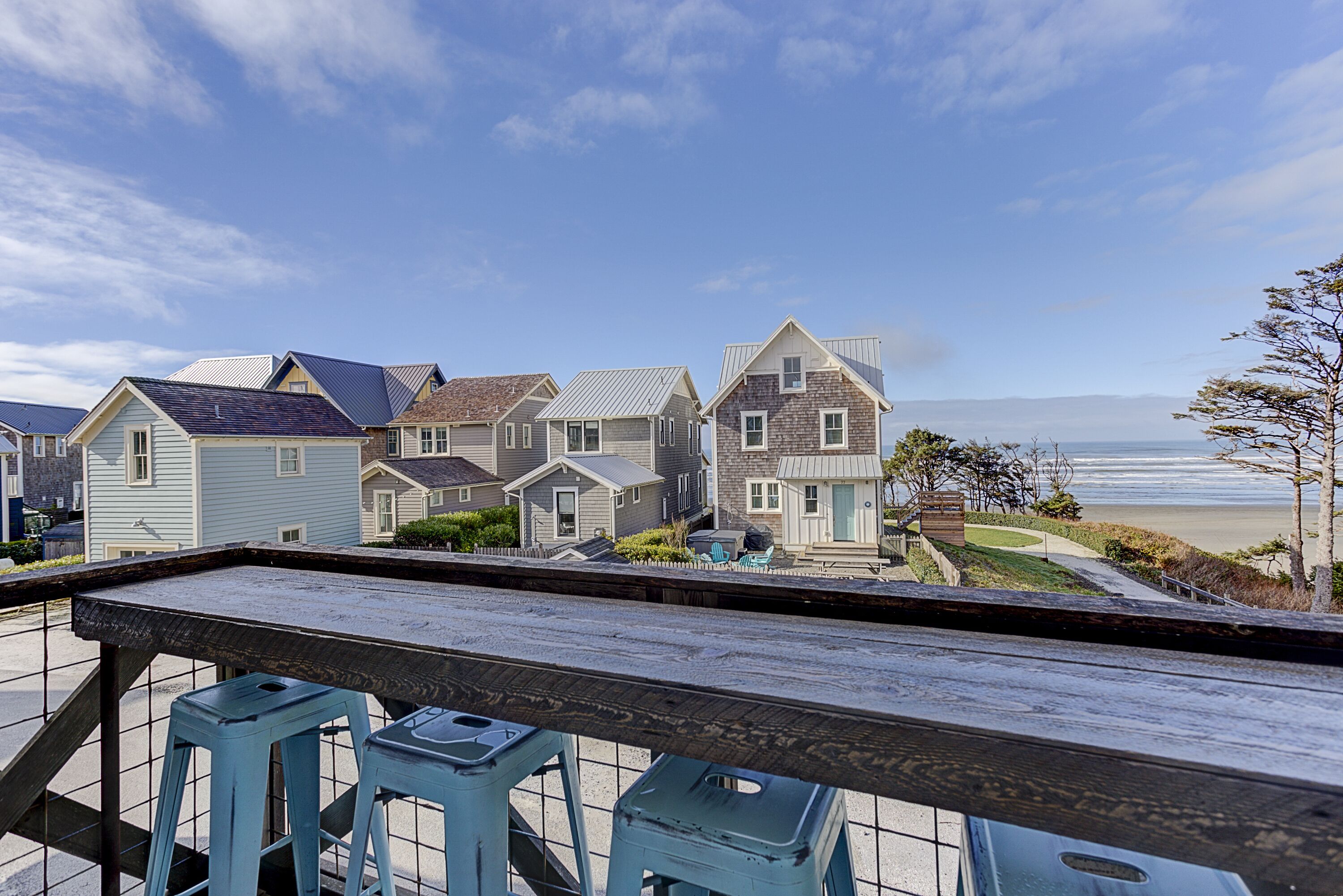 Une terrasse avec des tabourets bleus donnant sur une rangée de maisons, d’où l’on peut admirer la plage et l’océan.