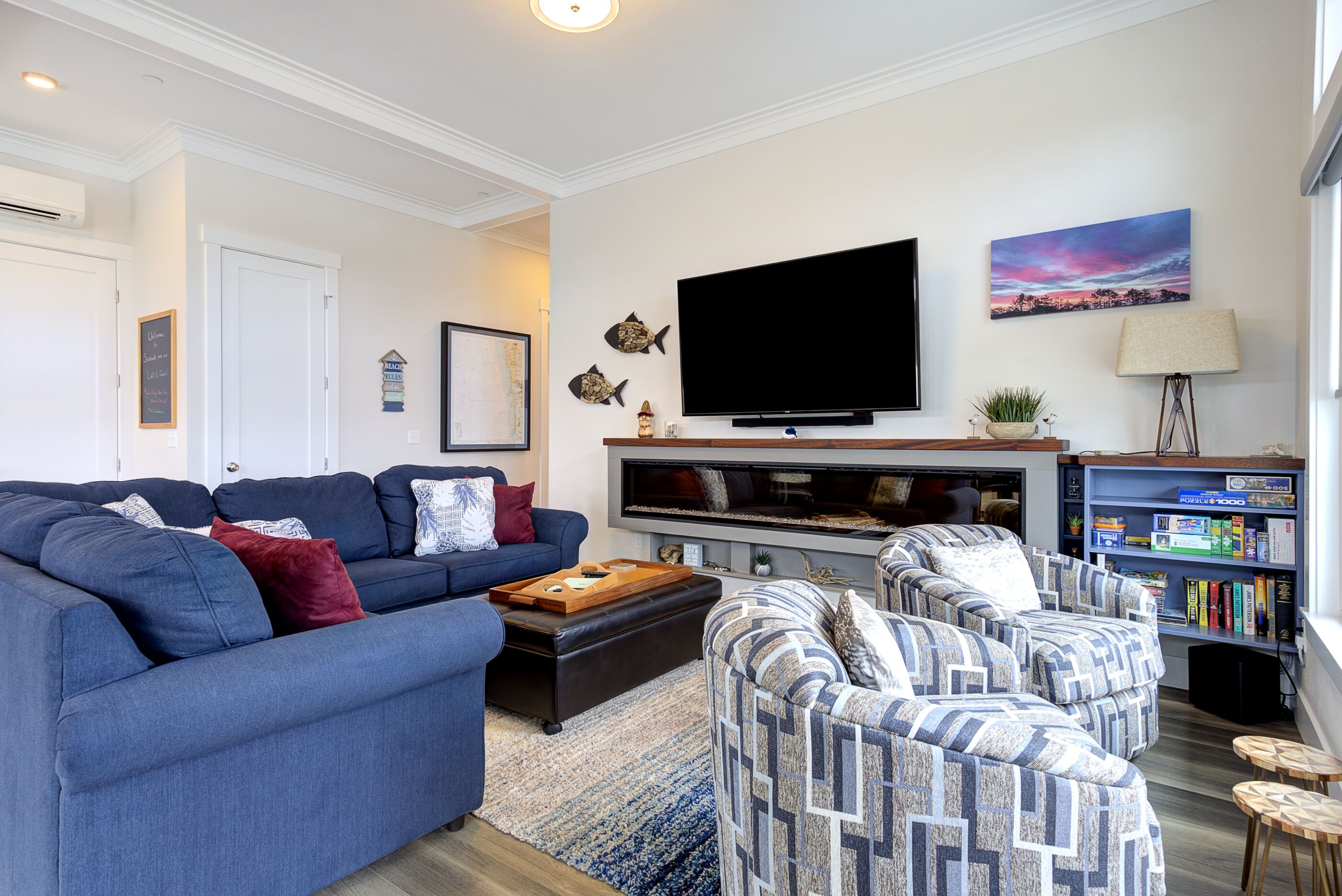 A modern living room with a blue sofa, patterned armchairs, a coffee table, a flat-screen TV, a bookshelf, and a painting on the wall.