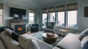 A living room with a fireplace, a television, a large window with a view of the beach, and a comfortable seating area. - Seabrook Love Shack | Oceanfront, Hot Tub, Ocean Views, Sleeps 6 (Pacific Beach)