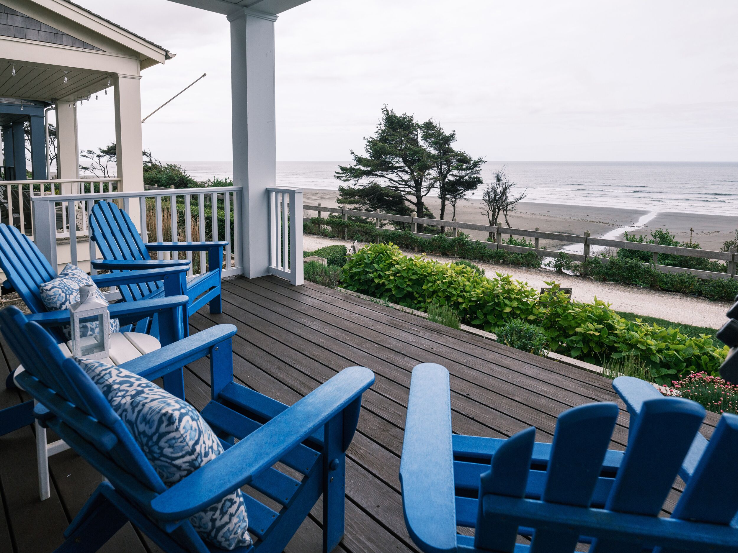Eine Holzterrasse mit blauen Adirondack-Stühlen und Blick auf den Strand.