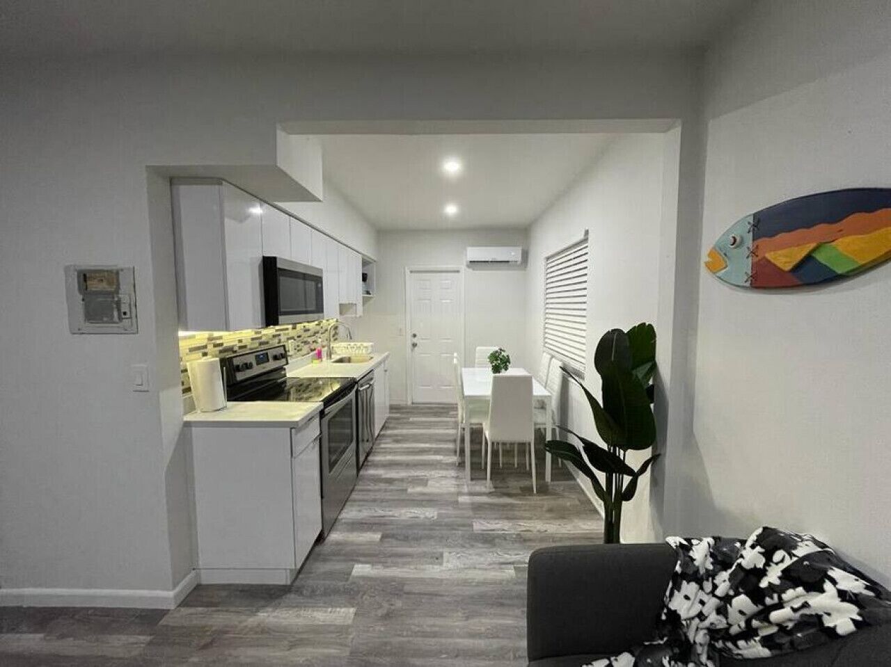 A modern kitchen with white cabinets, a dining area with a table and chairs, and a living room with a sofa and a plant.
