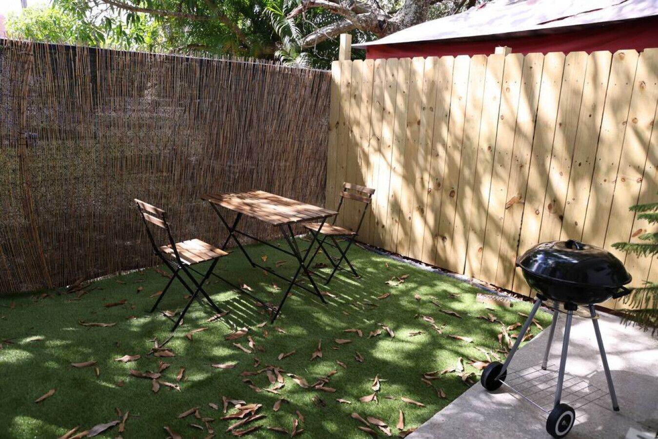Un jardin avec une table et des chaises en bois, un barbecue et une clôture en bambou.