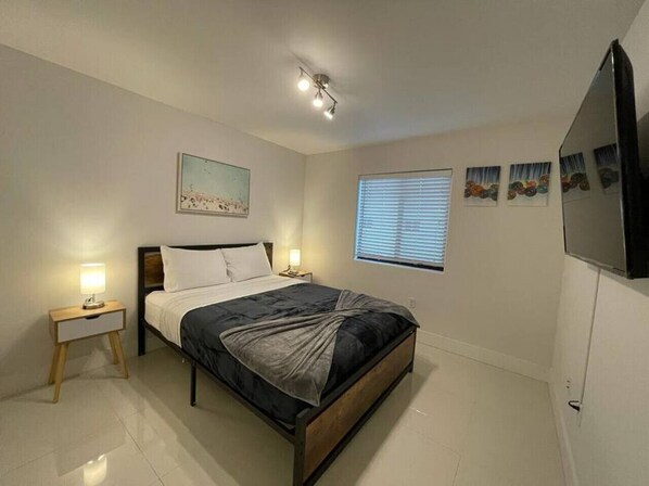Un dormitorio con cama, mesitas de noche, luz de techo, ventana con persianas y decoración en la pared.