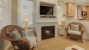 A living room with a fireplace, two wicker chairs, a television mounted above the fireplace, and wooden cabinets in the adjacent kitchen area. - Seabrook Ocean Eyrie | Pet Friendly, Fire Pit | 2BR, Sleeps 5, EV Charging (Pacific Beach)