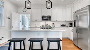 A modern kitchen with white cabinetry, a central island with bar stools, a stainless steel refrigerator, and a microwave. - Seabrook Oceanview Home | Dog Friendly | Hot Tub, Pool & Gym | Sleeps 12 (Pacific Beach)