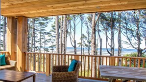 Een houten terras met rieten meubilair, met uitzicht op een strand en hoge dennenbomen.