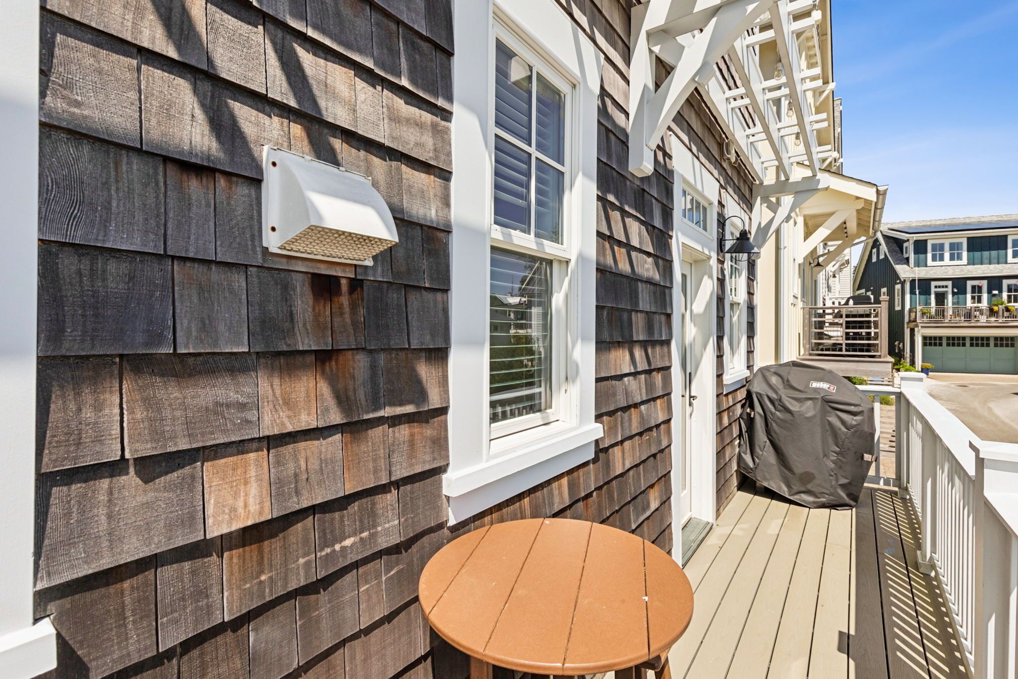 Une terrasse en bois avec une table ronde, un barbecue couvert et un mur extérieur blanc agrémenté d’une fenêtre.