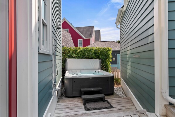 Un bain à remous installé dans un espace extérieur étroit, agrémenté d’une terrasse en bois, et entouré de maisons dont les murs sont recouverts de bardage vert et rouge.