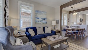 Ein Wohnzimmer mit blauem Sofa, einem grauen Sessel, einem Couchtisch aus Holz und einem Essbereich mit Holztisch und Stühlen.