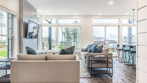 Sala de estar moderna com sofá, mesa de centro e televisão na parede. Grandes janelas oferecem vista para o exterior.