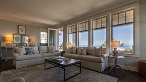 A spacious living room with a large window offering a view of the ocean, a beige sofa set, a coffee table, and two floor lamps. - Seabrook Sandy Side Up | Oceanfront, Hot Tub | 4BR, Sleeps 10, Seastack Views (Pacific Beach)