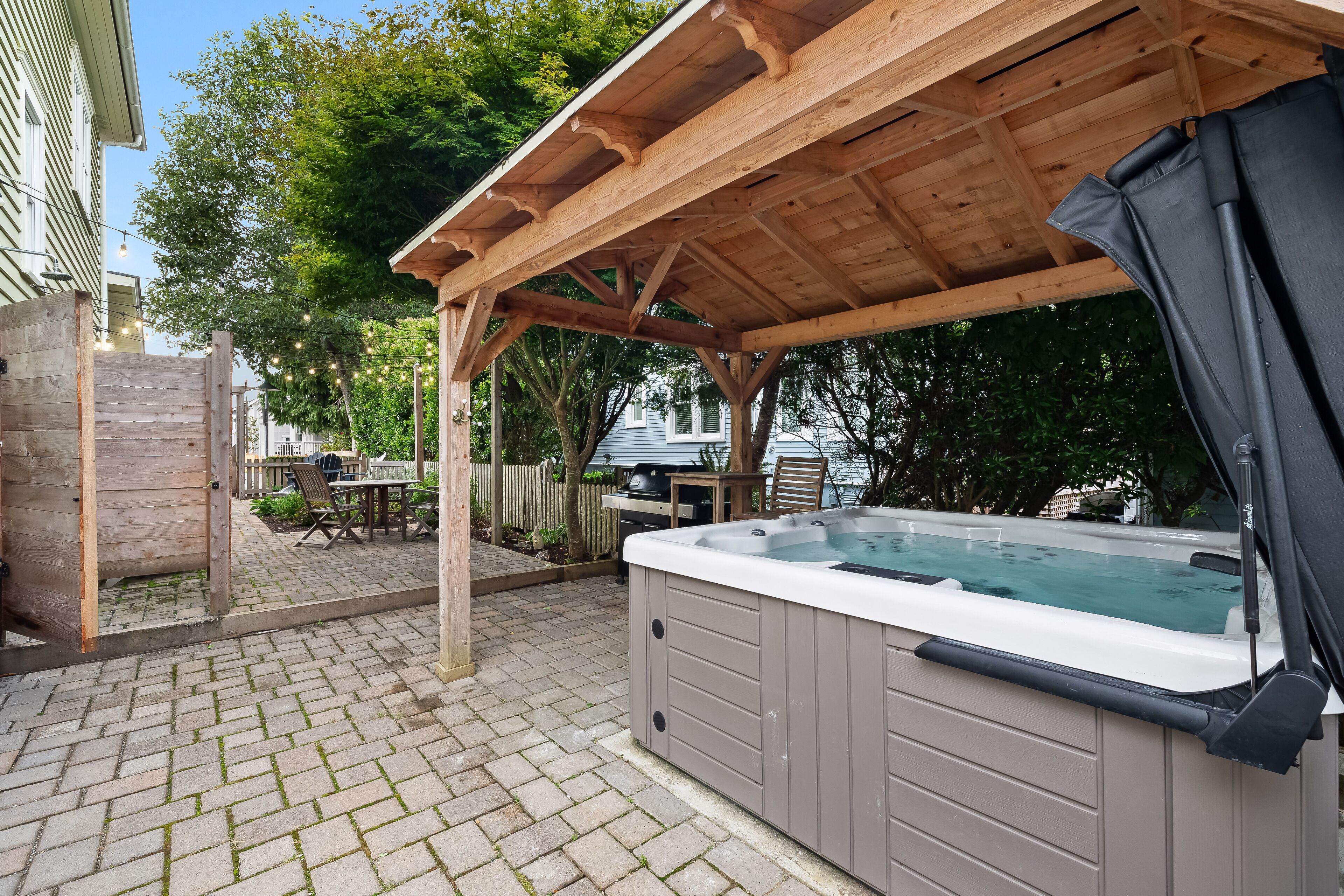 A hot tub under a wooden pergola in a backyard with a brick patio, surrounded by trees and a fence.