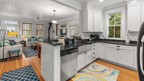 A modern kitchen with white cabinetry, a black countertop, and a built-in dishwasher. There is a dining area with a table and chairs, and a living room with a sofa and a lamp.