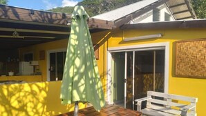 Une maison jaune avec une table et un banc en bois, un parasol vert et une porte-fenêtre coulissante.