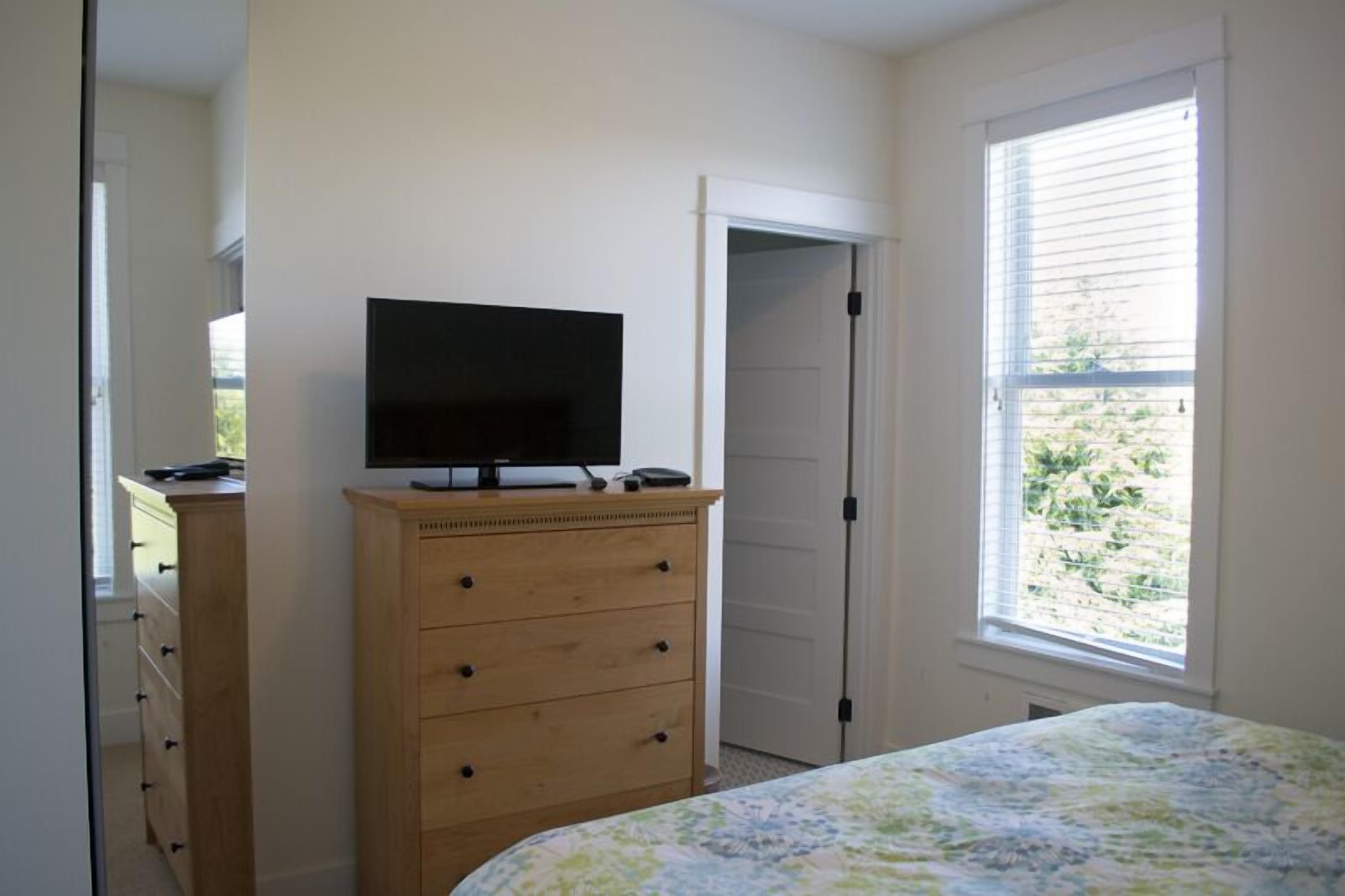Ein Schlafzimmer mit Bett, Schminktisch, Fernseher auf dem Schminktisch, einer Tür und einem Fenster mit Jalousien.