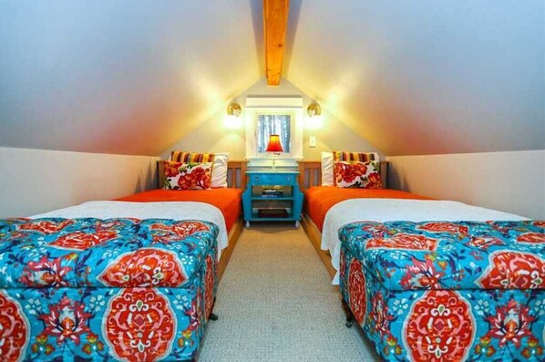 A room with two beds, each with colorful patterned bedding, a small table with a lamp, and a window with curtains.